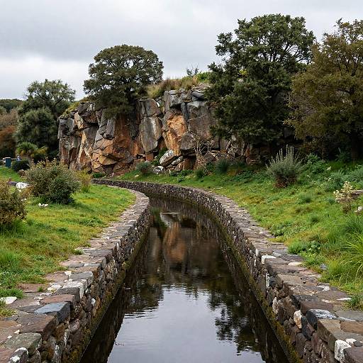 Photograph of a narrow, stone-bordered canal reflecting rocky cliffs and lush greenery under a cloudy sky, with trees in the background.