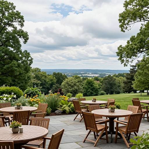 Photograph of a scenic outdoor café with wooden tables and chairs, surrounded by lush greenery and colorful flowers, overlooking a distant landscape under a cloudy sky
