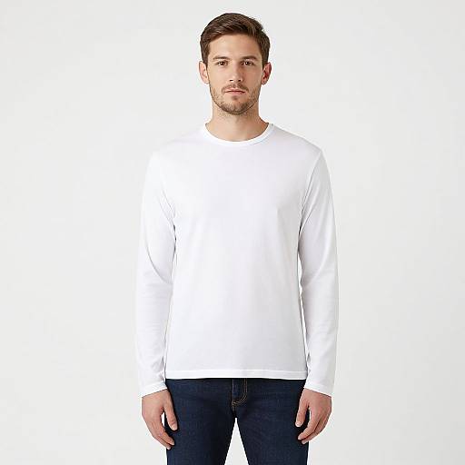 Photograph of a serious, young Caucasian man with short brown hair, wearing a plain white long-sleeve shirt and dark blue jeans, standing against