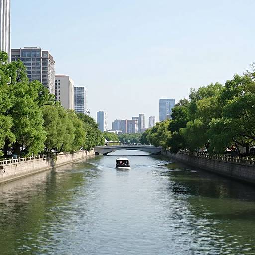 Photograph of a serene urban river with a small boat, flanked by lush green trees and tall modern buildings under a bright blue sky.