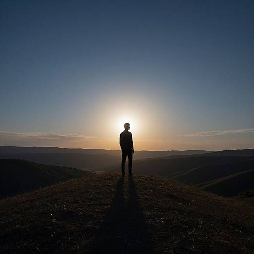Silhouetted hiker stands on rocky mountain peak at sunrise, casting long shadow, under clear blue sky with faint clouds.