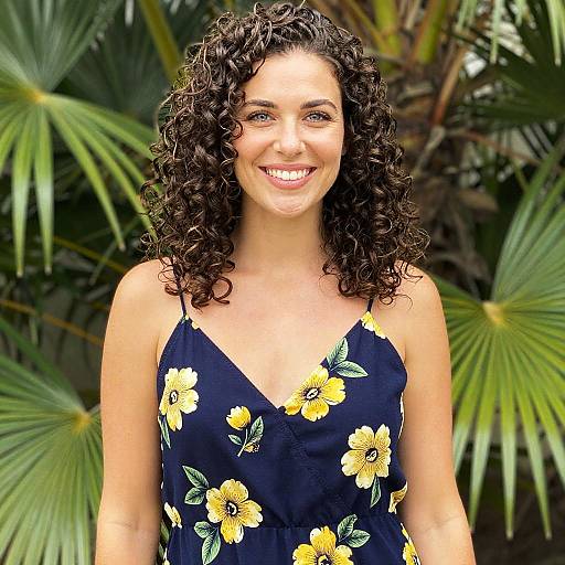 Photograph of a smiling woman with curly brown hair, wearing a black floral sundress, standing in front of lush green palm leaves.