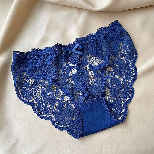 Photograph of vibrant blue lace panties with intricate floral patterns, lying on smooth, cream-colored satin fabric. The lace's delicate texture contrasts with the satin