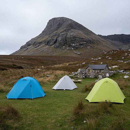 Photograph of three colorful tents (blue, white, yellow) in a grassy field with a rocky mountain and stone house in the background. Over