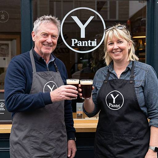 Photograph of smiling middle-aged man and woman, both in black Panii aprons, holding coffee cups, standing in front of a cafe window with