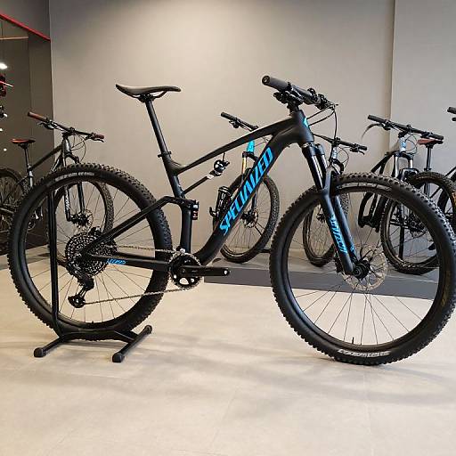 Photograph of a black Specialized mountain bike with blue accents, displayed on a stand in a well-lit showroom. Multiple bikes in the background.