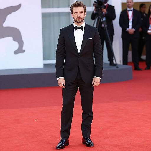 Photograph of a handsome man with dark hair and beard, wearing a black tuxedo and bowtie, standing on a red carpet. Background includes