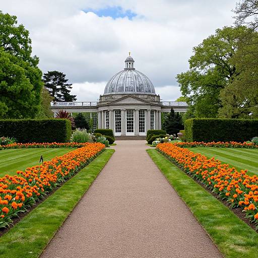 Photograph of a symmetrical garden with a central gravel path flanked by vibrant orange flowers, leading to a grand glass-domed building under a cloudy