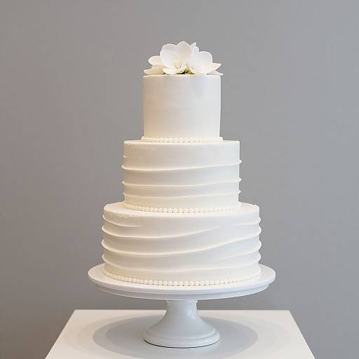 Photograph of a white, three-tiered, ruffled cake with small dot accents, topped by a white flower, on a white pedestal stand against