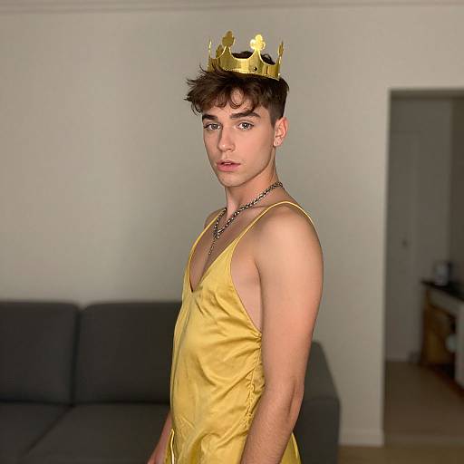 Photograph of a young, androgynous person with fair skin, brown hair, wearing a gold crown, yellow satin tank top, and black