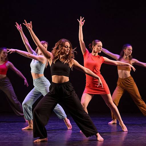 Photograph of five dancers in mid-performance, wearing colorful, form-fitting tops and loose pants, arms raised, against a black background.