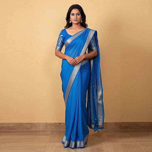 Photograph of a dark-haired woman in a vibrant blue saree with silver embellishments, standing against a beige wall, hands clasped.