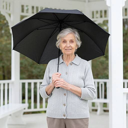 Photograph of an elderly woman with short gray hair, wearing a light gray knit cardigan and holding a black umbrella, standing on a white porch with