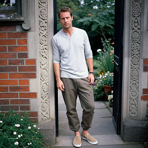 Photograph of a handsome, fair-skinned man with short brown hair, wearing a white long-sleeve henley, gray pants, and beige