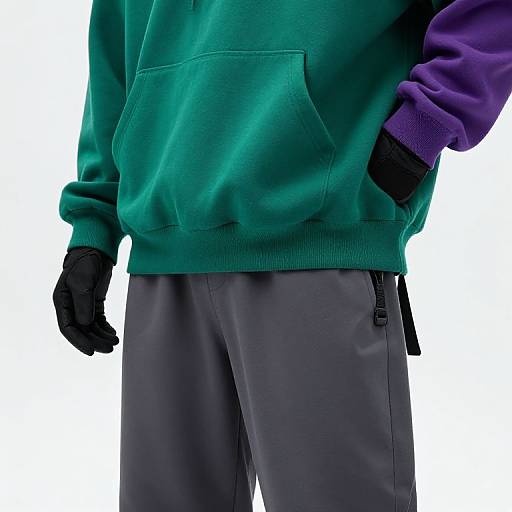 Photograph of a person wearing a green hoodie with purple sleeves, black gloves, and gray pants, hands in pockets, white background.