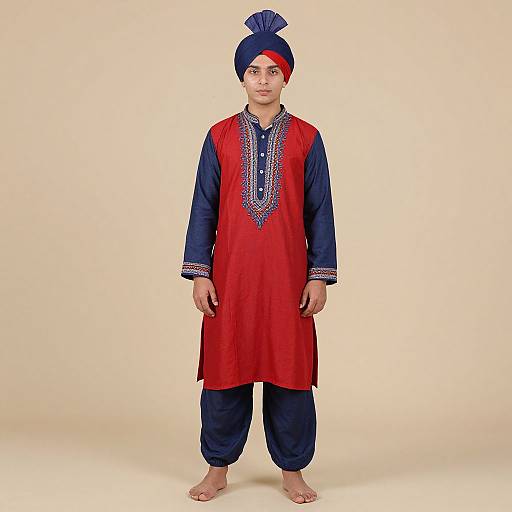 Photograph of a young boy in traditional Punjabi attire: red kurta with blue sleeves, intricate embroidery, navy trousers, barefoot, and a