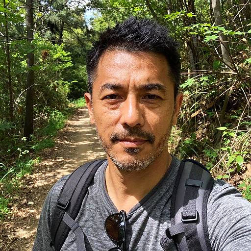 Photograph of an Asian man with short black hair and goatee, wearing a gray t-shirt, black backpack, and sunglasses, standing on a sunny