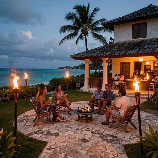 Photograph of sunset gathering: five adults, diverse ethnicities, in casual summer clothes, sitting around a fire pit on a stone patio, tropical house