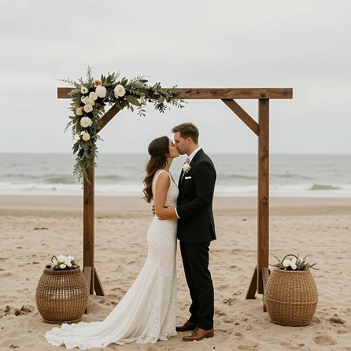Photograph of a bride and groom kissing at a beach wedding, standing under a wooden arch adorned with flowers, with wicker baskets on either side.