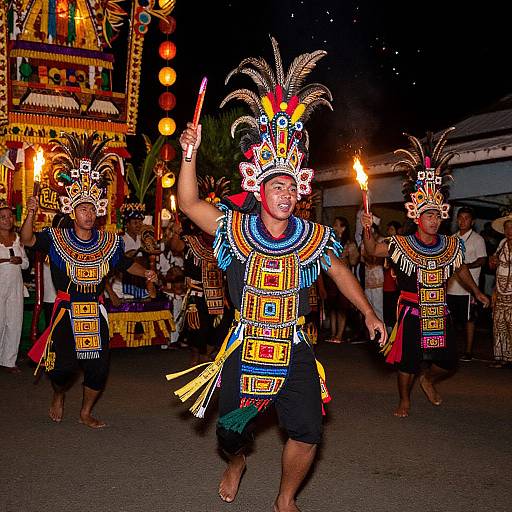 Photograph of three male dancers in colorful, intricate indigenous costumes with feathered headdresses, raising hands and torches, performing at night.