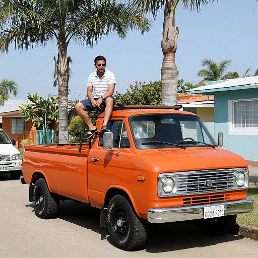 Photograph of a man in a white shirt and sunglasses sitting on the bed of an orange vintage pickup truck, parked on a sunny street with palm trees