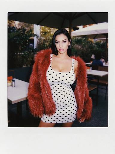 Photograph of a pregnant woman with dark hair, wearing a white polka dot dress and a red fur jacket, standing outdoors at a restaurant.
