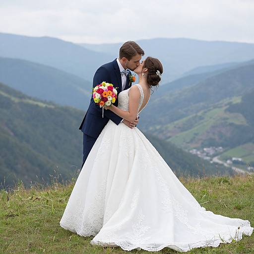 Photograph of a bride in a white lace gown and groom in a navy suit kissing on a grassy mountain peak, with a colorful bouquet and h