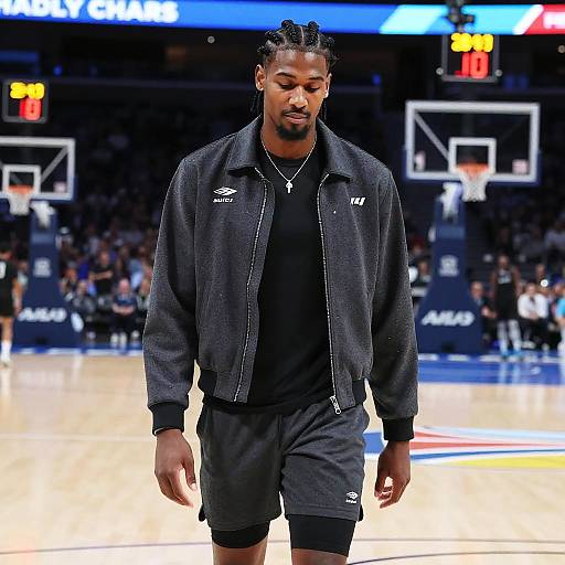 Photograph of a tall, muscular Black male basketball player with braided hair, wearing a black jacket and shorts, walking on a brightly lit basketball court