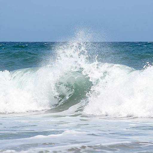 Photograph of a powerful ocean wave crashing, with white frothy water splashing against a dark blue sea under a clear sky.
