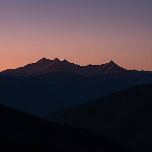 Photograph of a mountain silhouette at sunset, featuring a gradient sky from pink to purple, with dark, shadowed mountain peaks in the foreground.