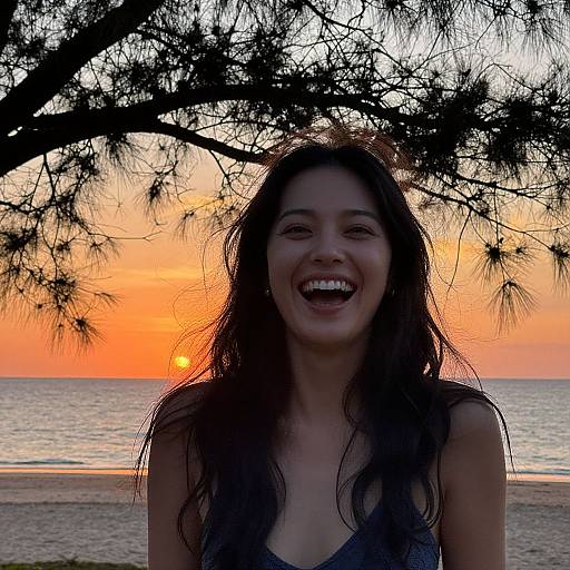 Photograph of a smiling young woman with long, dark hair, standing at a beach during sunset, silhouetted by tree branches, with an