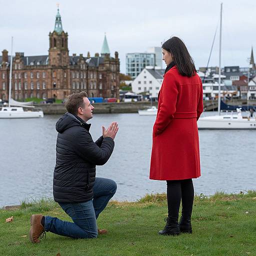 Photograph of a bearded man kneeling, hands clasped, proposing to a woman in a red coat standing by a waterfront with historic buildings and boats