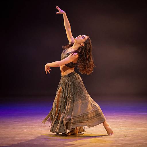 Photograph of a dark-haired dancer in a black beaded crop top and flowing skirt, elegantly posing with arms raised against a dark, illuminated stage