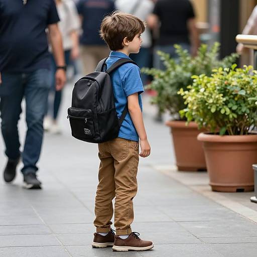 Photograph of a young boy with brown hair, blue shirt, tan pants, brown shoes, black backpack, standing on a city sidewalk. Blurred