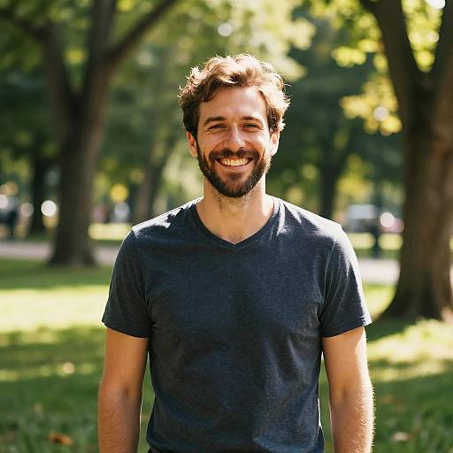 Photograph of a smiling bearded man with short brown hair, wearing a dark blue V-neck T-shirt, standing in a sunlit park with trees