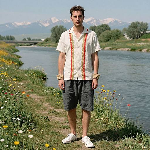 Photograph of a young man with short dark hair, wearing a white shirt, black shorts, white shoes, and beaded necklaces, standing by