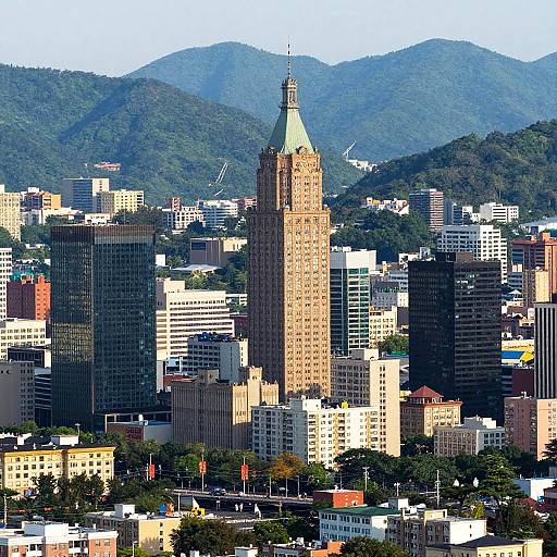 Photograph of a city skyline with a tall, tan skyscraper center, surrounded by various buildings, and green mountainous background under a clear blue sky