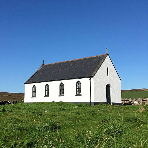 Photograph of a simple white church with dark roof, arched windows, and grassy hill background under a clear blue sky.