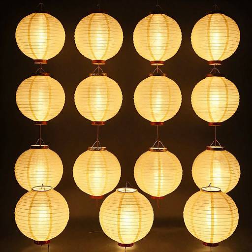 Photograph of twelve glowing, spherical, yellow paper lanterns arranged in a 3x4 grid against a dark background, creating a warm, illuminated