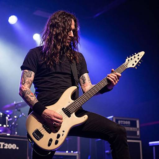 Photograph of a tattooed, long-haired male guitarist in black shirt and pants, playing a cream-colored electric guitar on stage under blue and white lights