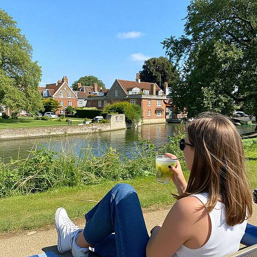 Photograph of a woman with brown hair, wearing a white tank top and blue jeans, sitting by a river, drinking a yellow drink, with red