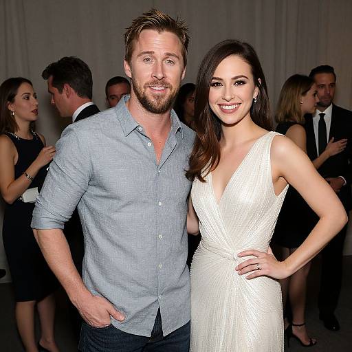Photograph of a bearded, blue-shirted man with brown hair, and a smiling woman in a white, deep V-neck dress, standing closely
