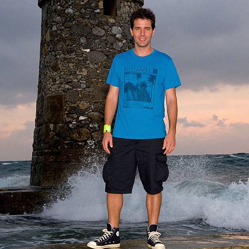 Photograph of a smiling man with short dark hair, wearing a blue t-shirt, black shorts, and black sneakers, standing in front of a stone