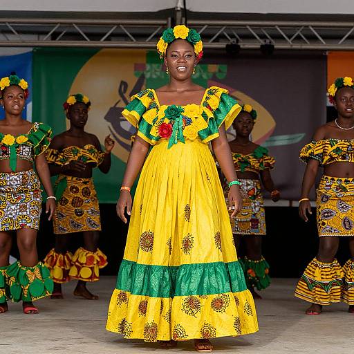 Photograph of a Black woman in a vibrant yellow and green traditional African dress with pineapple patterns, standing center stage with four dancers in similar outfits, decorated