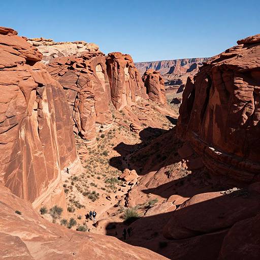 Photograph of a rugged, red rock canyon with steep, sunlit cliffs, sparse desert vegetation, and a bright blue sky overhead.