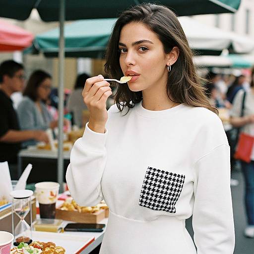Photograph of a young woman with long dark hair, wearing a white long-sleeve shirt with a black and white checkered patch, eating from