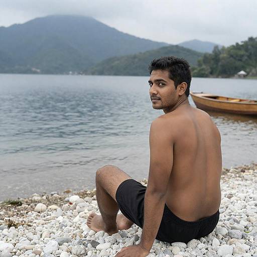 Shirtless Man Sitting on Pebbled Beach by Lake