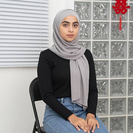 Photograph of a young woman with light brown skin, wearing a gray hijab, black long-sleeve top, and blue jeans, sitting on