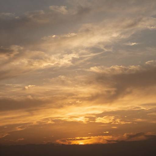 Photograph of a vibrant sunset sky, showcasing golden and orange hues with scattered clouds, and a dark silhouette of the horizon in the background.