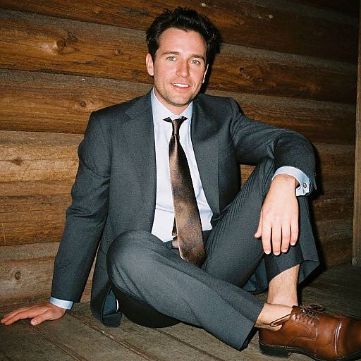 Photograph of a smiling, dark-haired man in a black suit, white shirt, brown tie, and brown shoes, sitting against a wooden log wall
